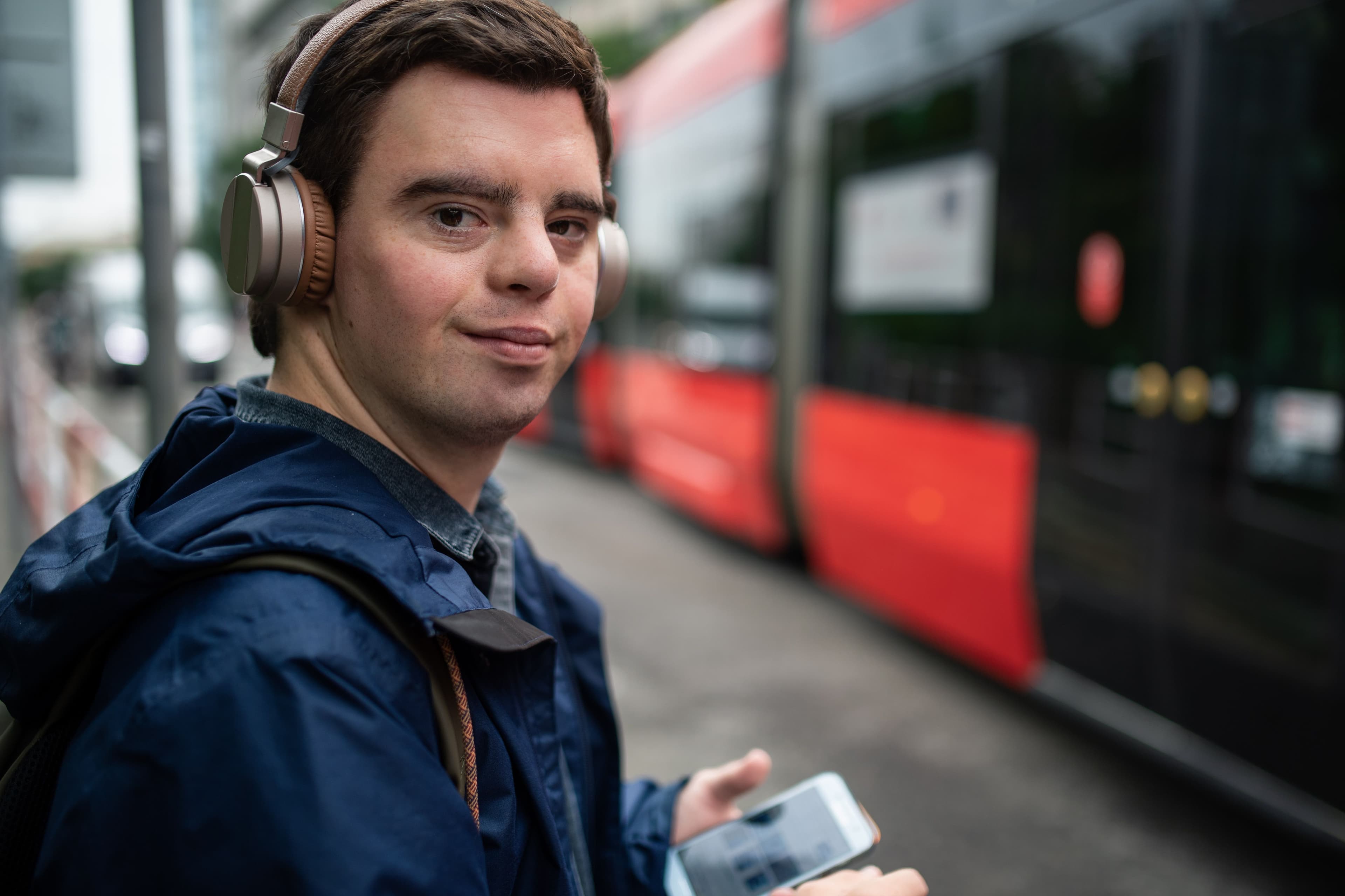 Mladý muž s Downovým syndrómom s nasadenými slúchadlami, ktorý drží v ruke tablet a stojí na zastávke, kde čaká na električku.